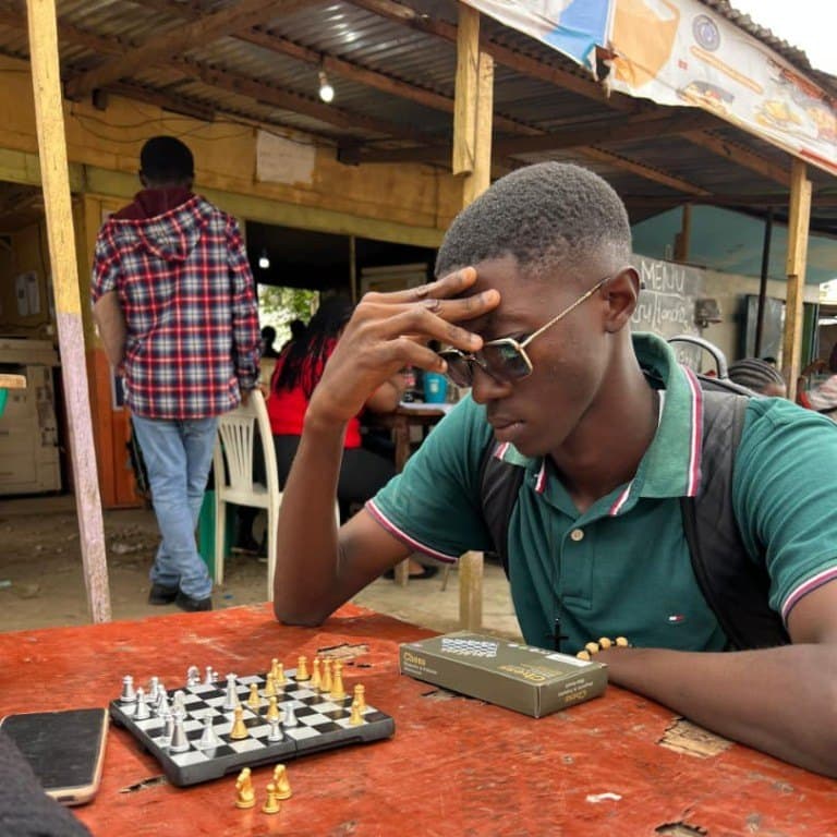 A student leans on one hand, thinking over a chess board on a table outdoors.
